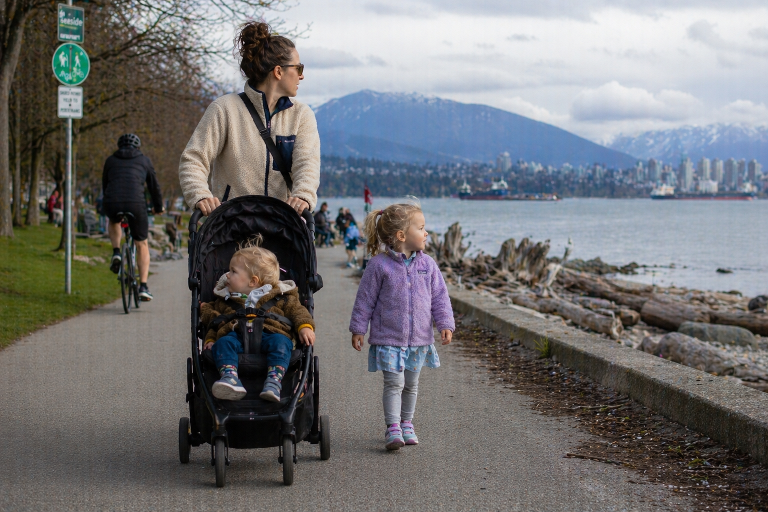 A family with two kids walking and running on the grass at a Vancouver seawall park, with mountains and the city skyline in the background.